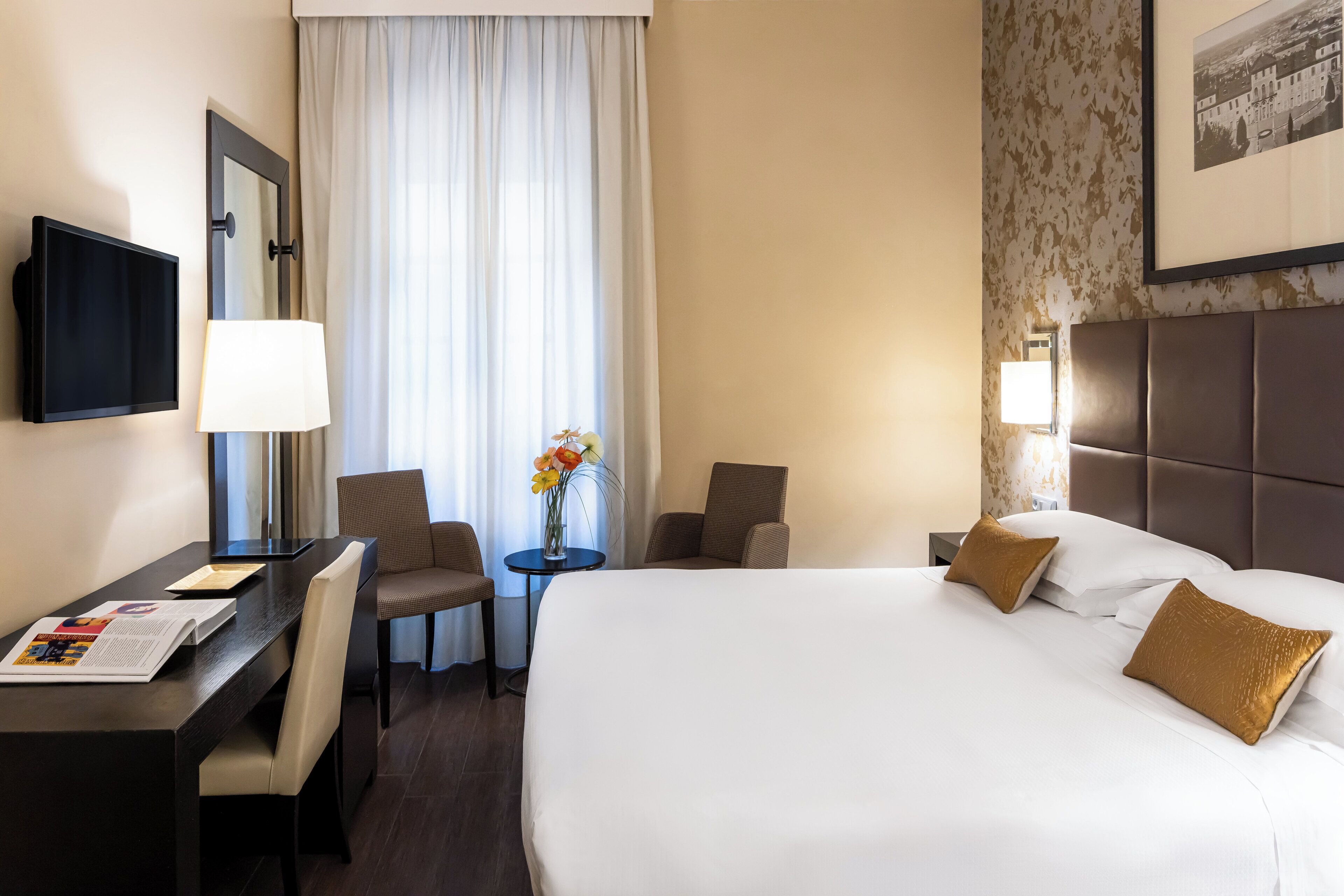 Una habitación de hotel con una cama grande, un escritorio con una silla, una mesita con un jarrón de flores y un televisor en la pared.
