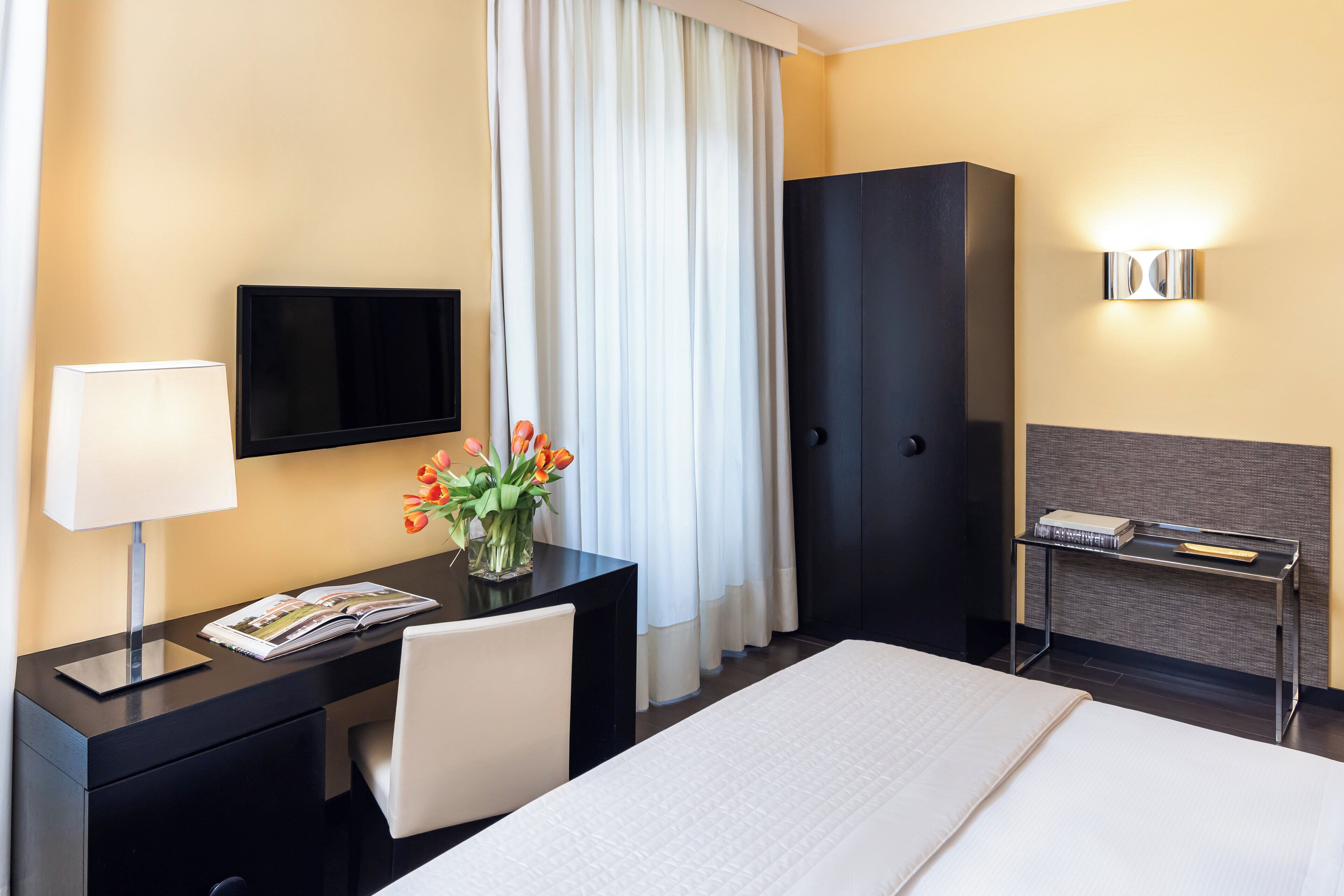 Habitación de hotel con cama, escritorio con lámpara, televisor, armario y mesita de noche con libros.