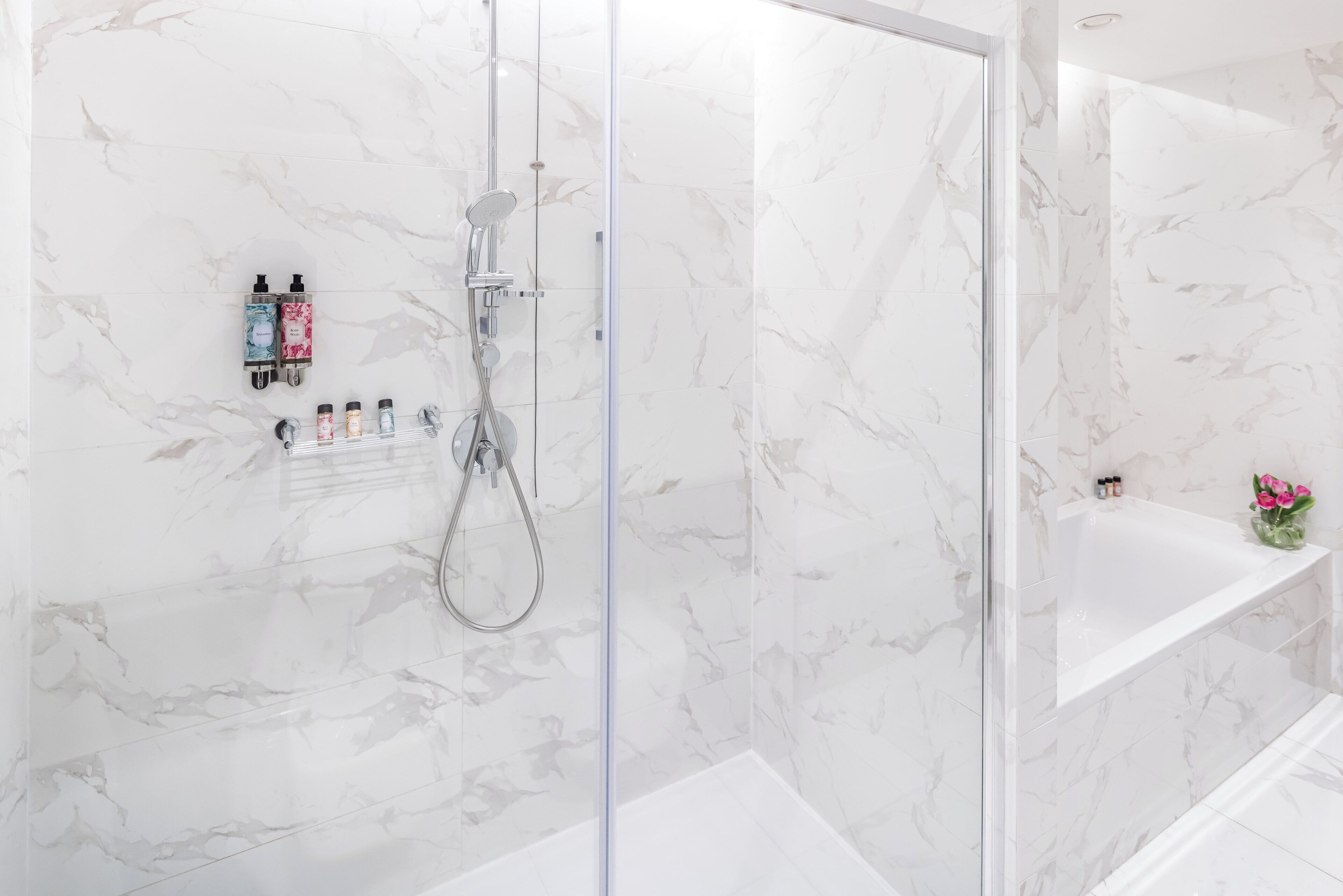 A modern bathroom with a glass-enclosed shower, marble walls, and a white bathtub with a shelf holding toiletries.