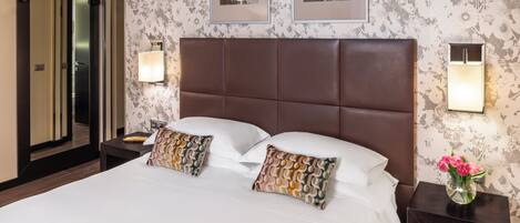 A hotel room with a large bed, two pillows, a nightstand with a vase of flowers, and framed pictures on the wall.