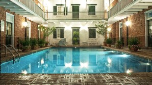 A swimming pool with a brick building in the background, a covered outdoor area, and potted plants around the pool. - Hotel Provincial (New Orleans)