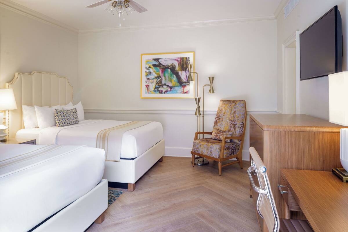 A hotel room with two beds, a desk, a chair, a TV, and a painting on the wall.