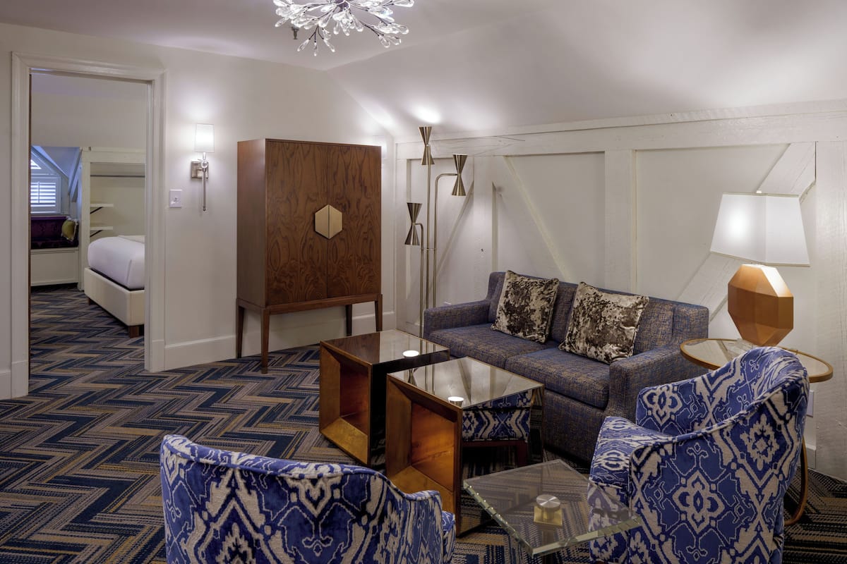 A hotel room with a sofa, two patterned armchairs, a glass coffee table, a wooden cabinet, and a bed in the background.