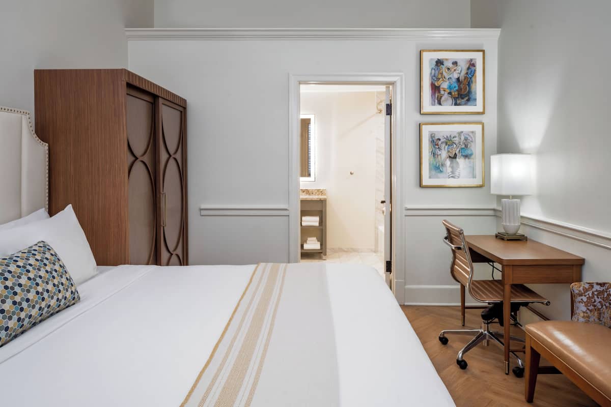 A hotel room with a large bed, a wooden wardrobe, a desk with a chair, and two framed pictures on the wall.