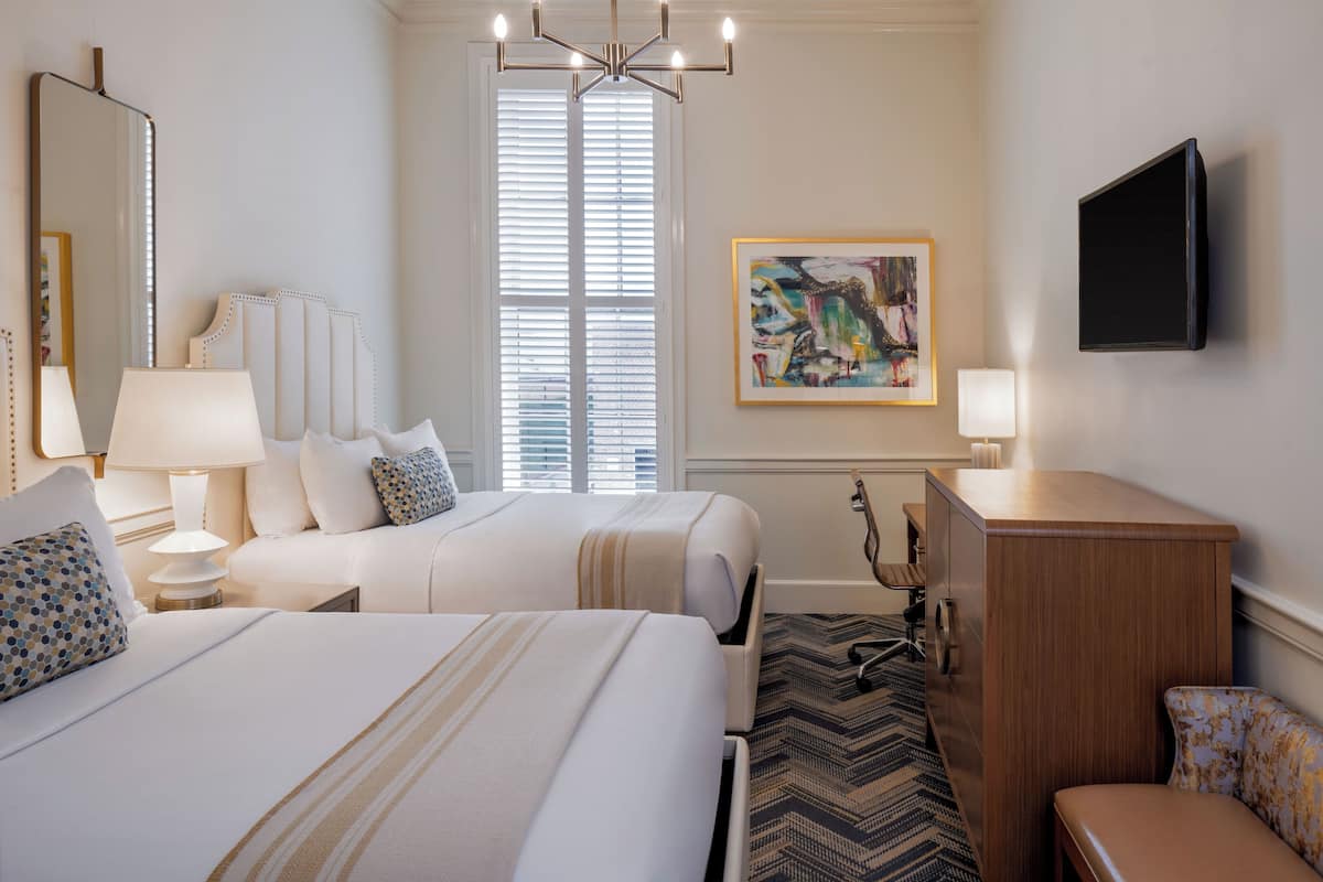 A hotel room with two beds, a desk, a chair, a TV, a painting, and a large window with blinds.