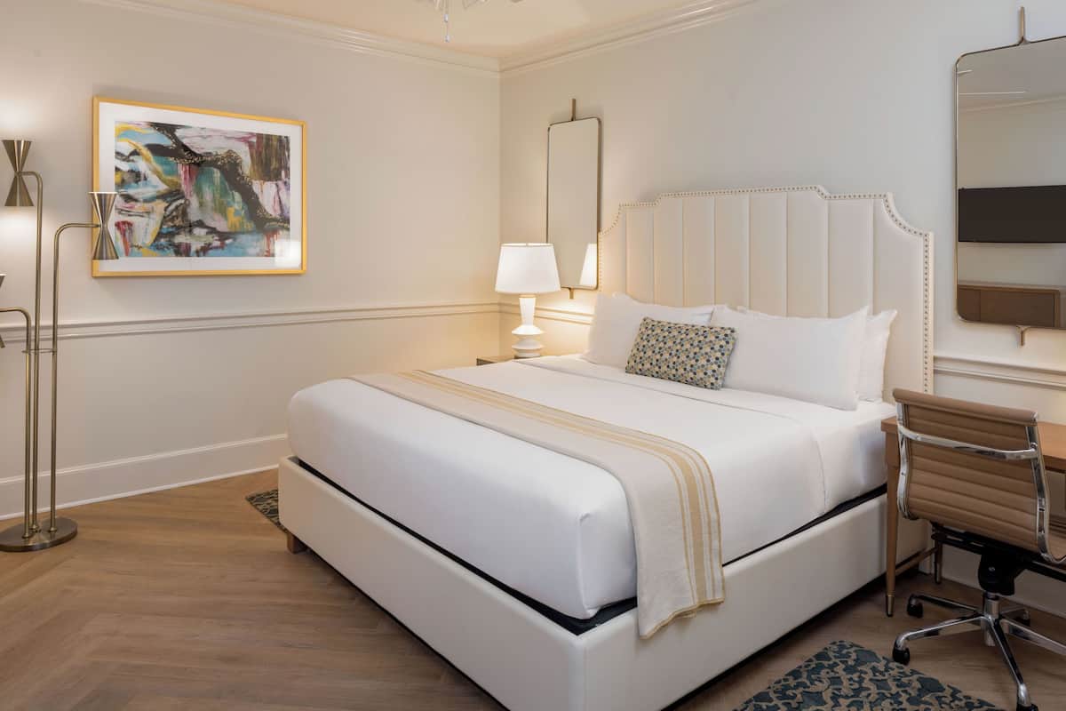 A hotel room with a large bed, a desk with a chair, a standing lamp, a painting on the wall, and a mirror.