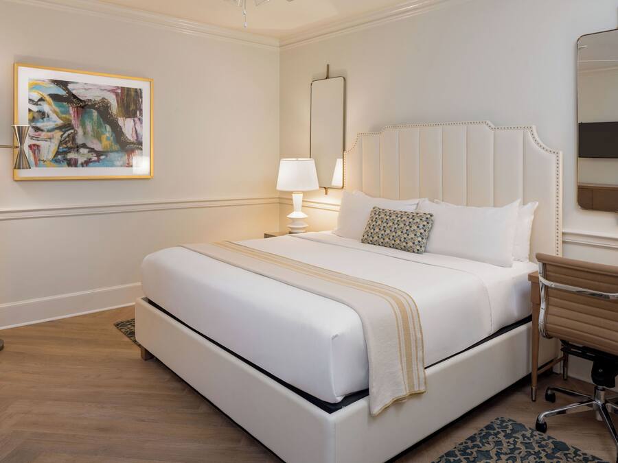 Habitación de hotel con una cama grande, un escritorio con silla, una lámpara de pie, un cuadro en la pared y un espejo.