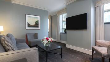 A living room with a grey sofa, a coffee table with a vase of flowers, a flat-screen TV mounted on the wall, and a patterned armchair.