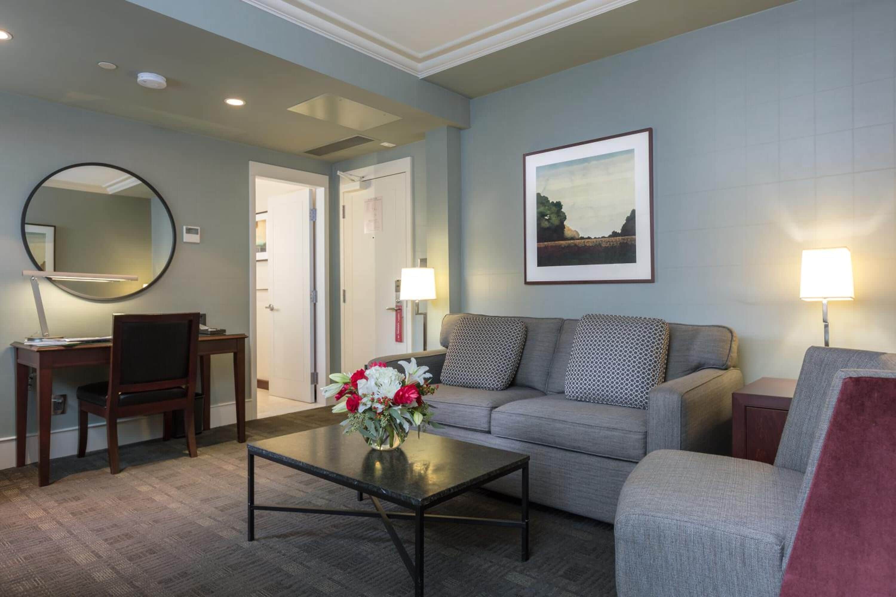A modern hotel room with a grey sofa, a round mirror, a desk with a chair, a coffee table with flowers, and a framed picture on the wall.