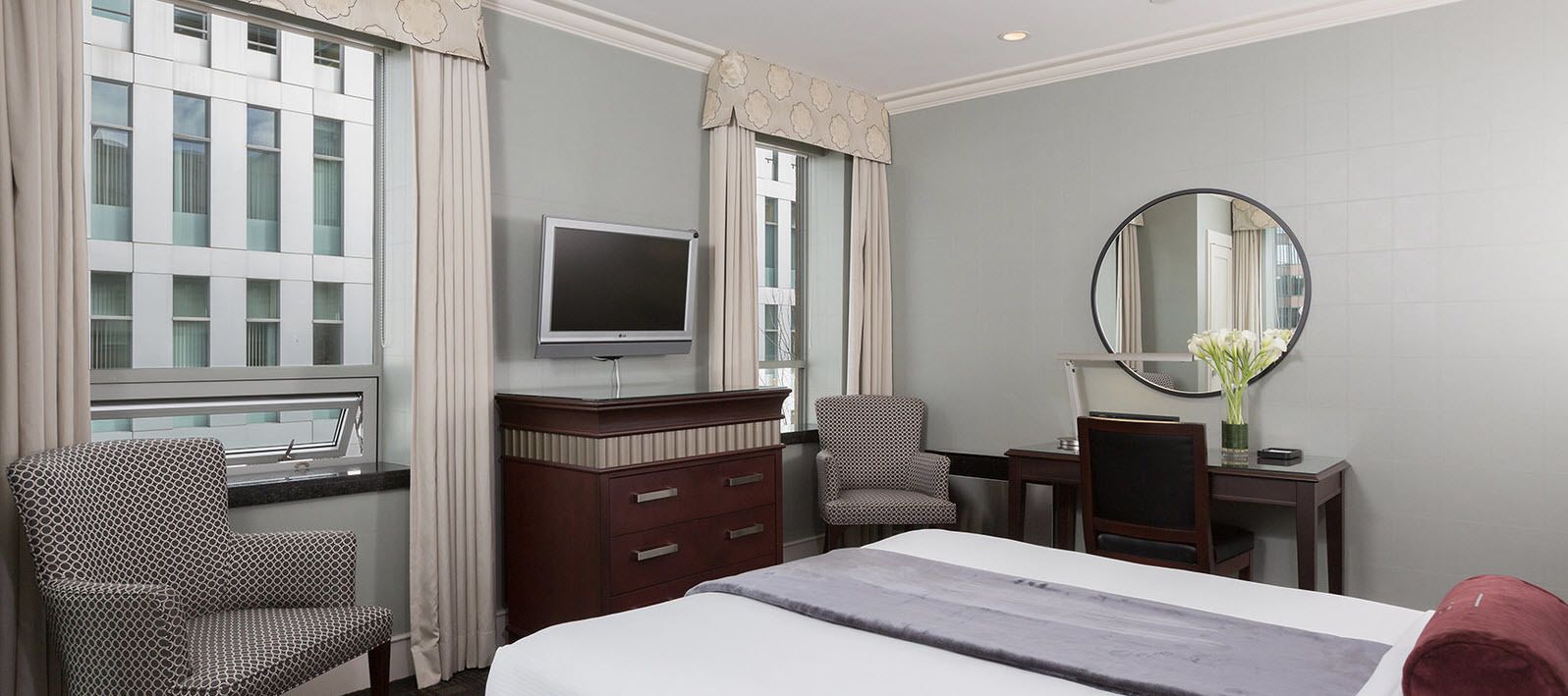 Una habitación de hotel con una cama, dos sillones estampados, un aparador, un televisor de pantalla plana, un espejo redondo y una ventana con cortinas.