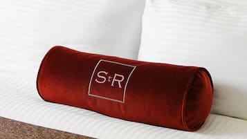 Un cojín de terciopelo rojo con un logotipo grabado sobre una cama con almohadas blancas y ropa de cama a rayas.
