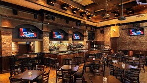 A restaurant with brick walls, multiple TVs displaying sports content, a bar area with a variety of bottles, and several tables and chairs for dining.