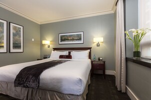 Habitación de hotel con una cama grande, dos mesitas de noche, un velador, un cuadro en la pared y una ventana con un jarrón de flores.