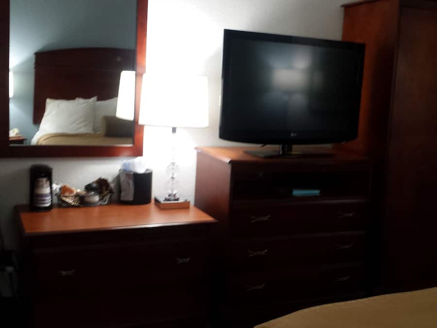 Quarto de hotel com cama, televisão em suporte de madeira, cômoda com gavetas, espelho e mesa de cabeceira com abajur e caixa de lenços.