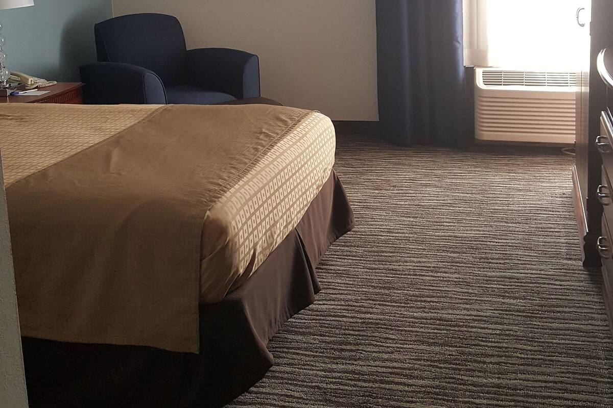 Quarto de hotel com uma cama, uma escrivaninha com abajur, uma cadeira, um quadro na parede e uma janela com cortinas.