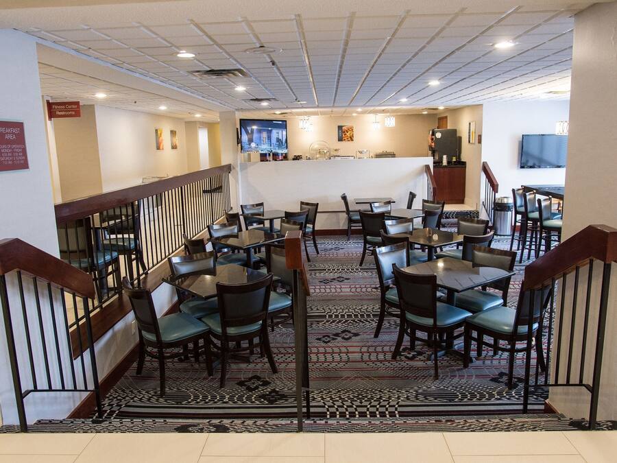 A hotel lobby with a staircase, dining area with tables and chairs, a fitness center sign, and a television screen.
