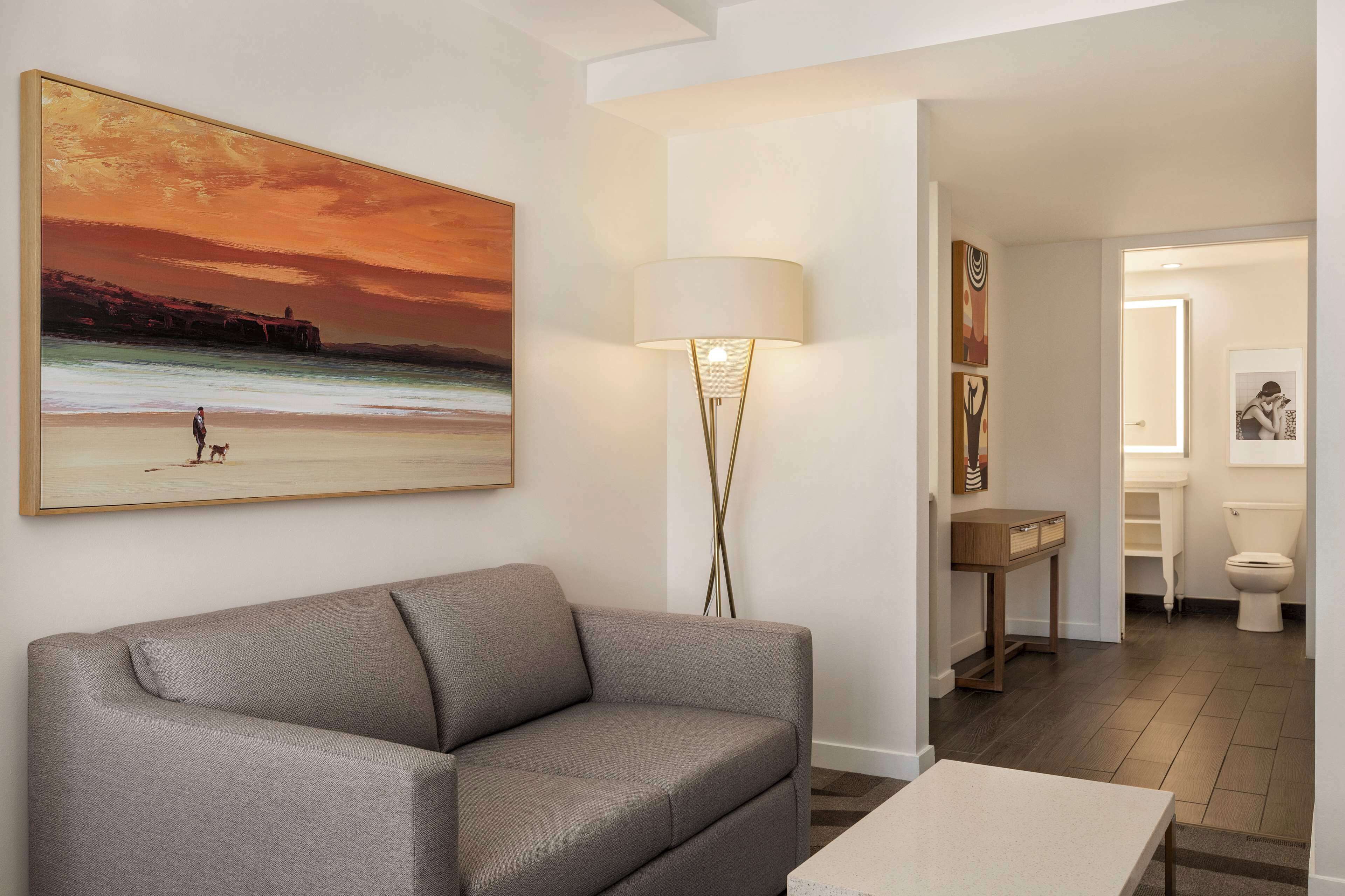 Un salon avec un canapé gris, un tableau représentant une scène de plage, une lampe sur pied et une vue sur la salle de bain.