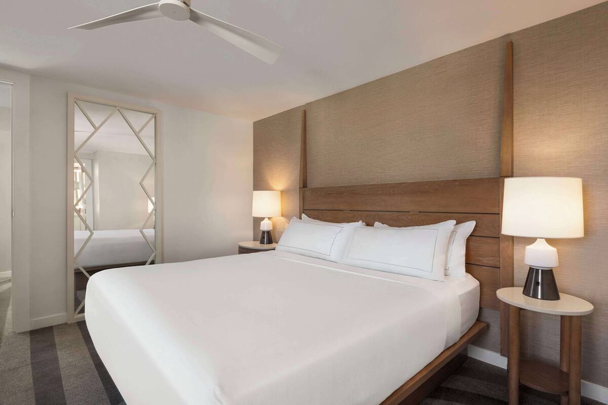 A hotel room with a large bed, two bedside tables with lamps, a ceiling fan, and a wooden headboard.