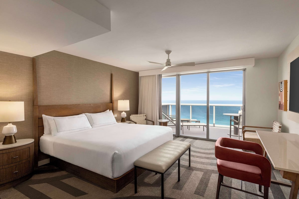 A hotel room with a large bed, a desk, a chair, and a balcony with a view of the ocean.
