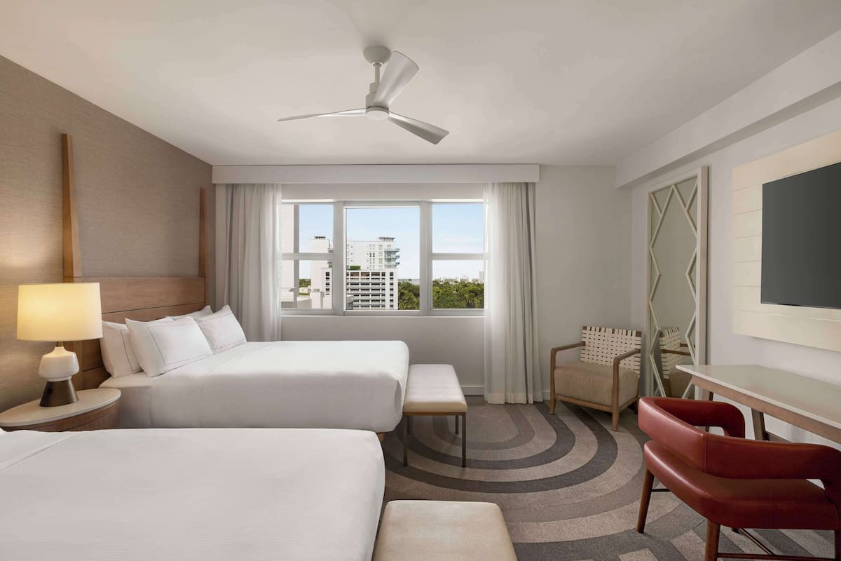 A hotel room with two beds, a desk, a chair, a TV, and a window with a city view.