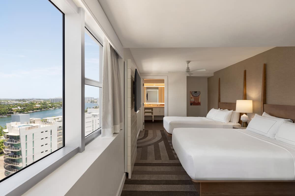 A hotel room with two beds, a large window with a city view, a bathroom, and a ceiling fan.