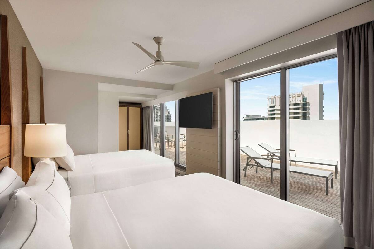 A hotel room with two beds, a ceiling fan, a TV, and a balcony with seating.