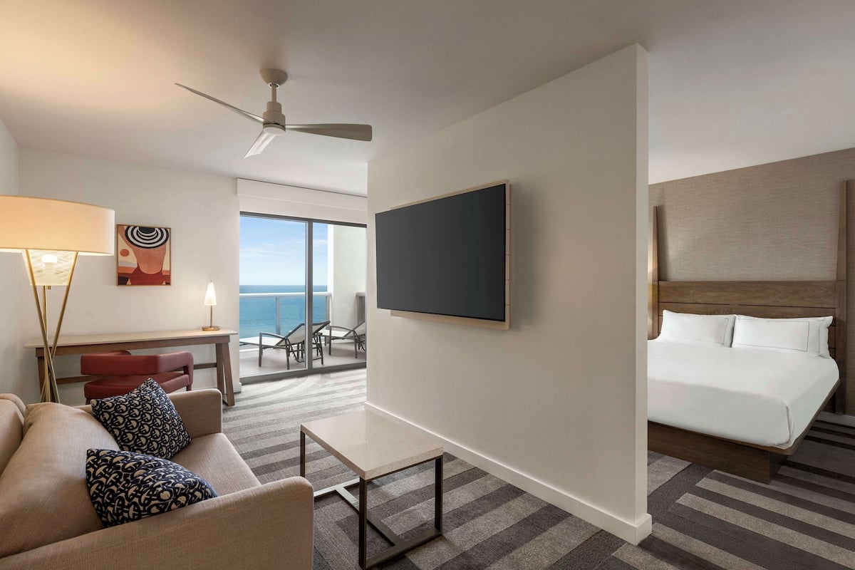 A hotel room with a bed, a TV, a desk, and a view of the ocean.