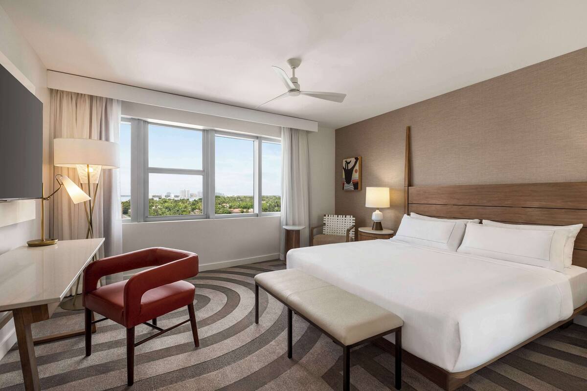 A hotel room with a large bed, a desk, a chair, a bench, a ceiling fan, and a window with a city view.