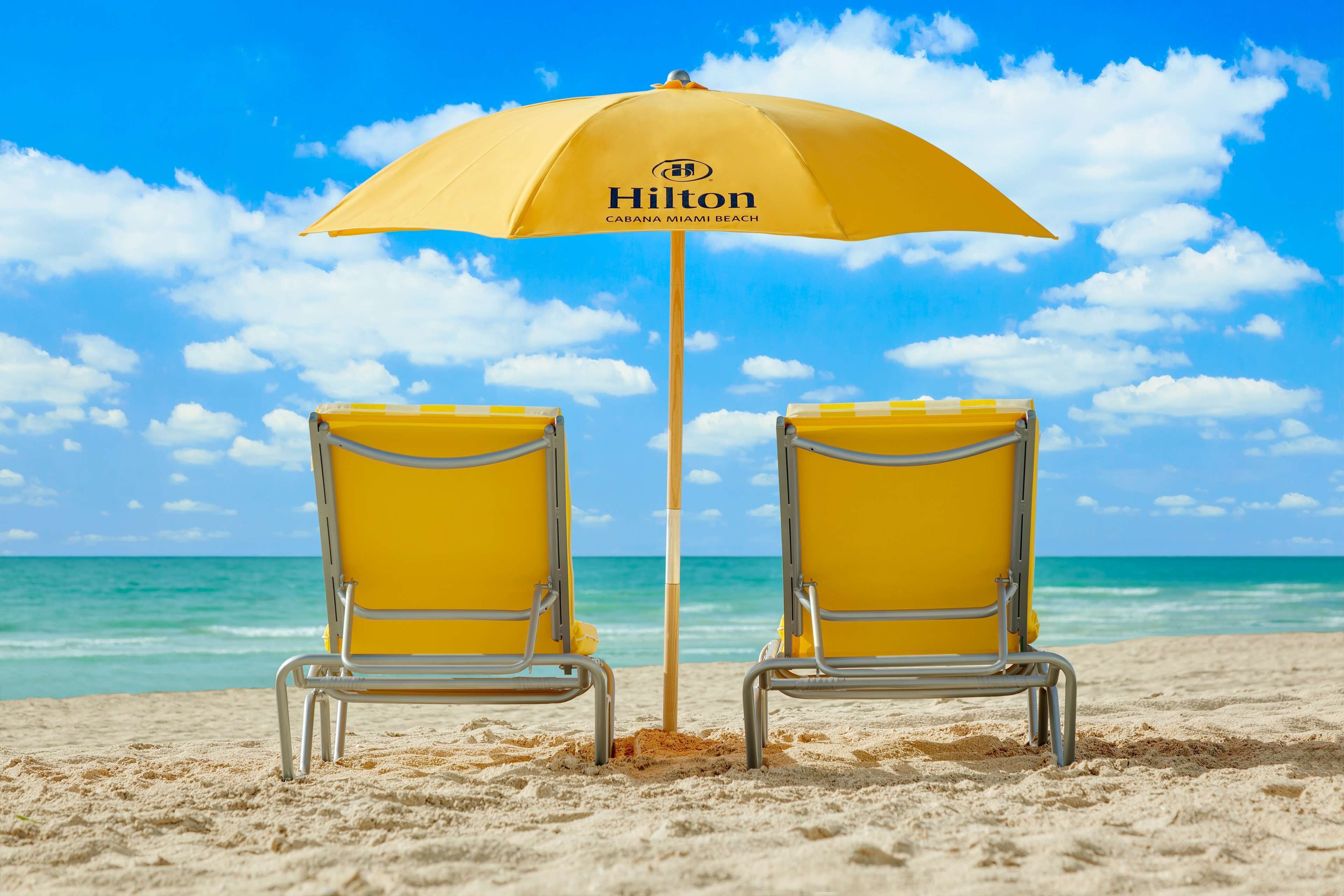 Twee gele strandstoelen onder een gele parasol met het Hilton logo, op een zandstrand met een helderblauwe lucht en oceaan op de achtergrond.