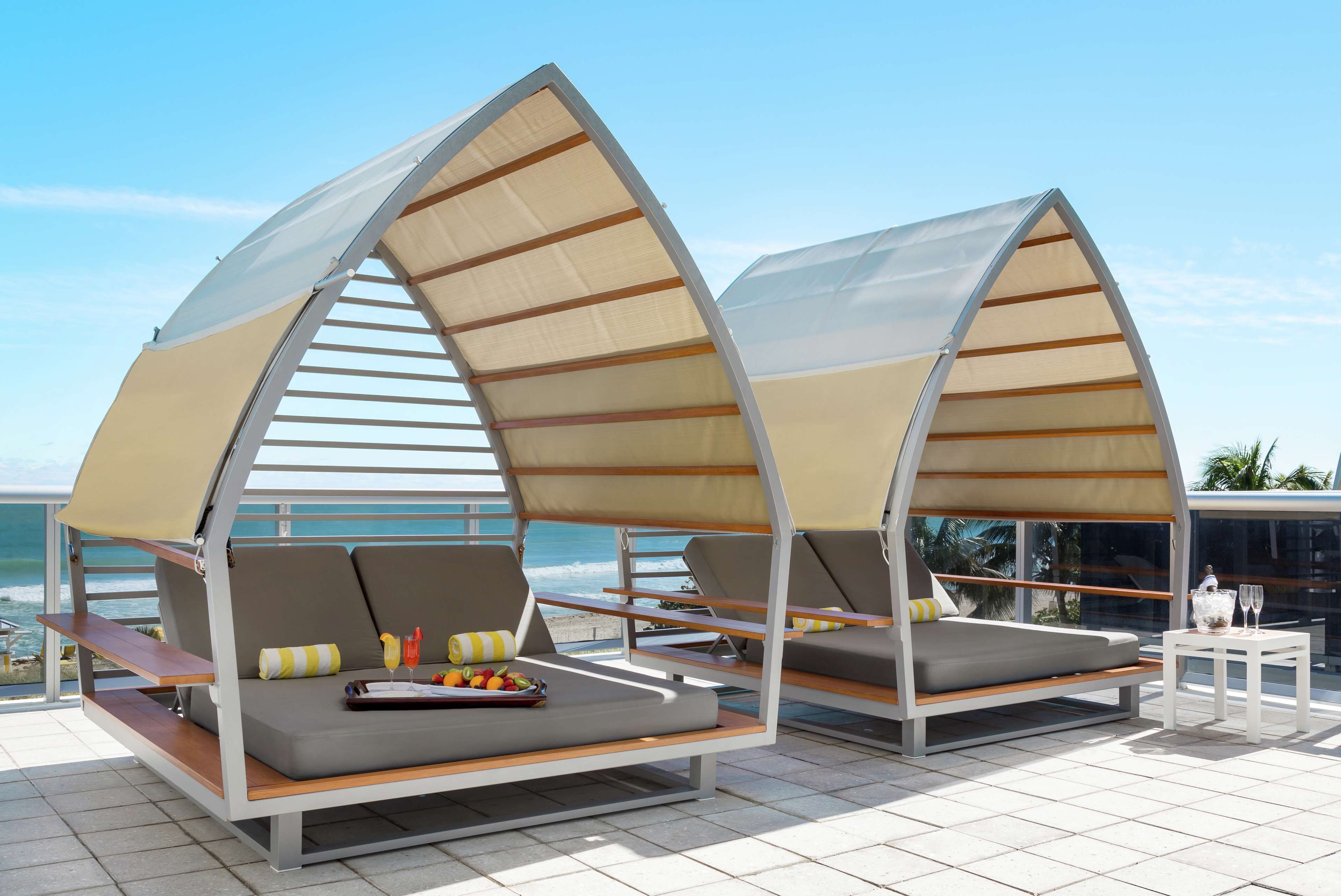 Two beachside lounge chairs with attached sunshades, a small table with a fruit bowl and glasses, and a view of the ocean.