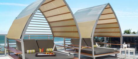 Two beachside lounge chairs with attached sunshades, a small table with a fruit bowl and glasses, and a view of the ocean.