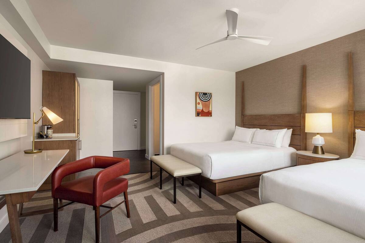 A hotel room with a bed, bedside tables, a desk, a chair, a bench, a ceiling fan, and a painting on the wall.