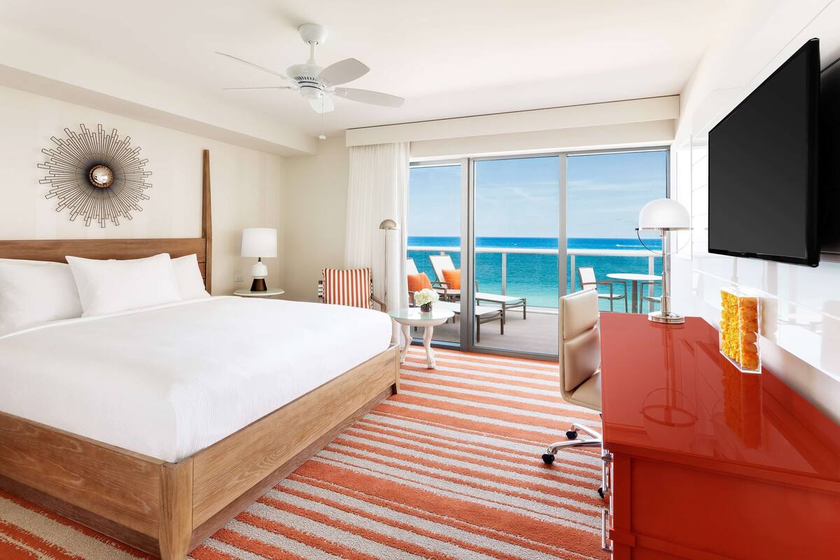 A hotel room with a bed, a desk, a TV, a ceiling fan, a large window with a view of the ocean, and a sunburst wall decoration.