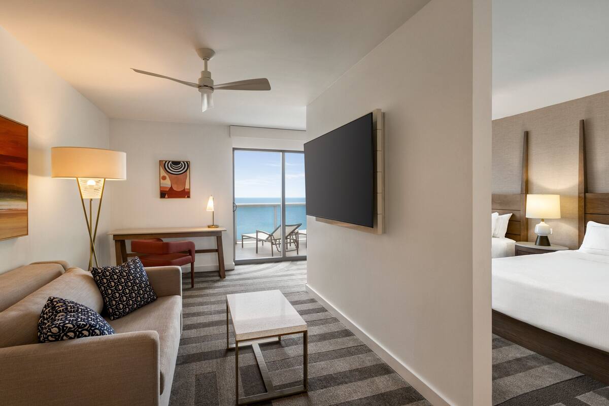 A hotel room with a bed, a sofa, a small table, a desk, a TV, a ceiling fan, and a view of the ocean.
