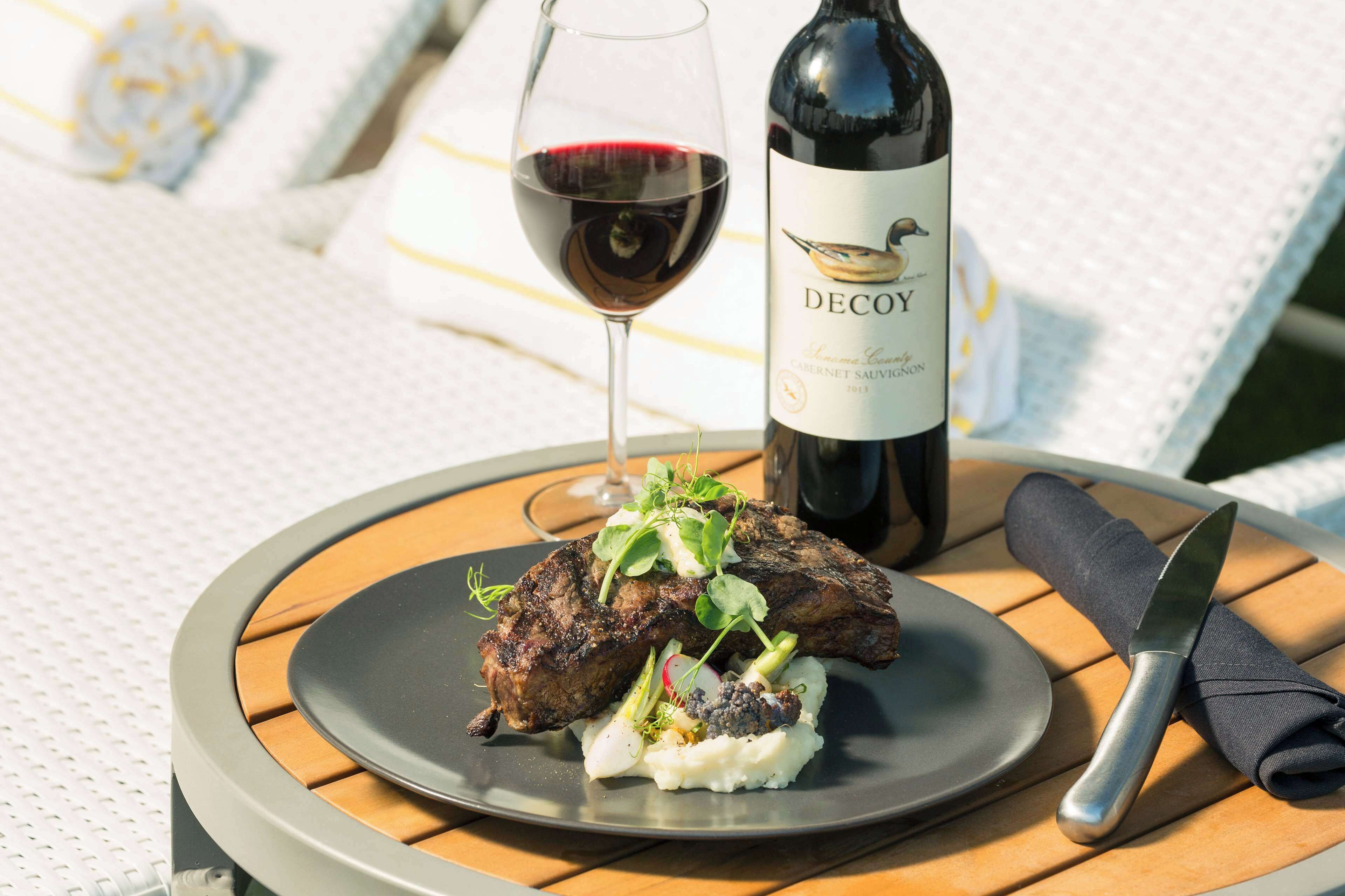 A plate with steak and mashed potatoes, a wine bottle, and a glass of red wine on a round table.