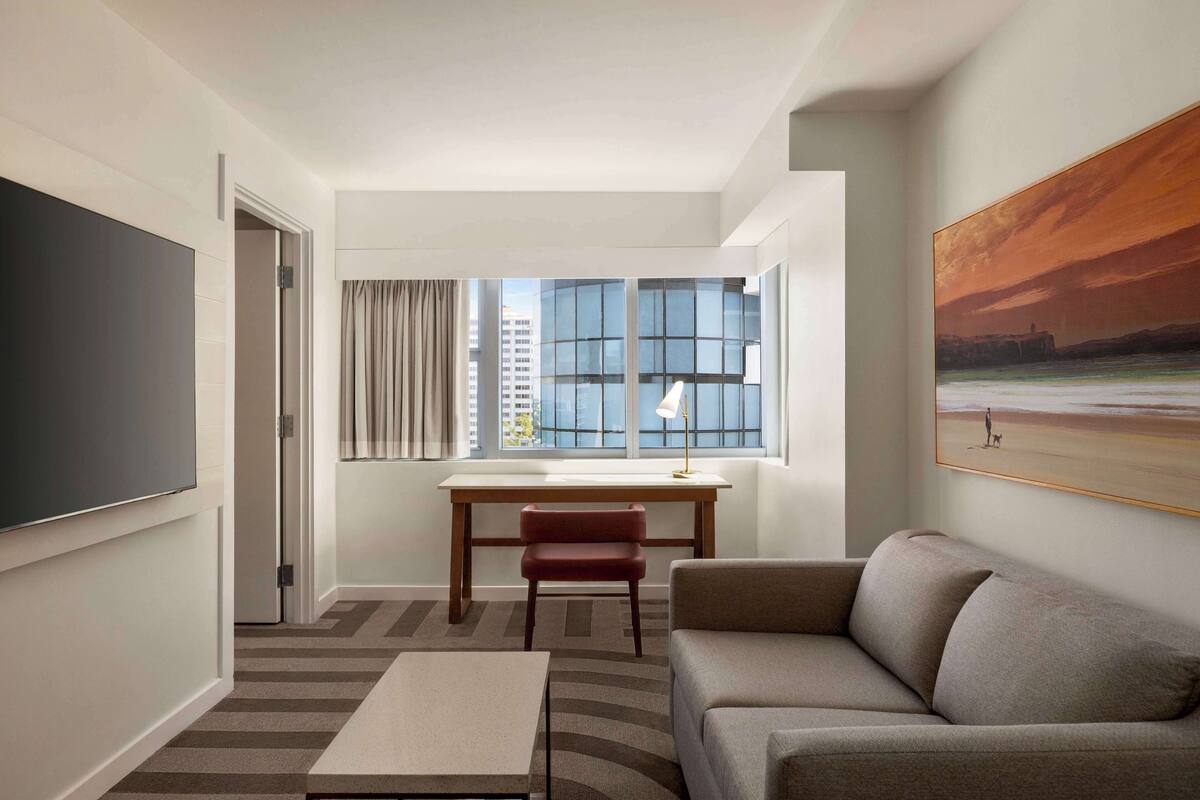 A modern hotel room with a large bed, a desk, a chair, and a view of the city through large windows.