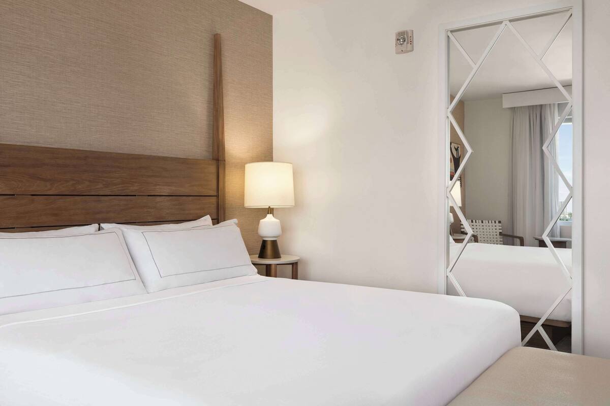 A hotel room with a large bed, a nightstand with a lamp, and a mirror with a geometric frame.