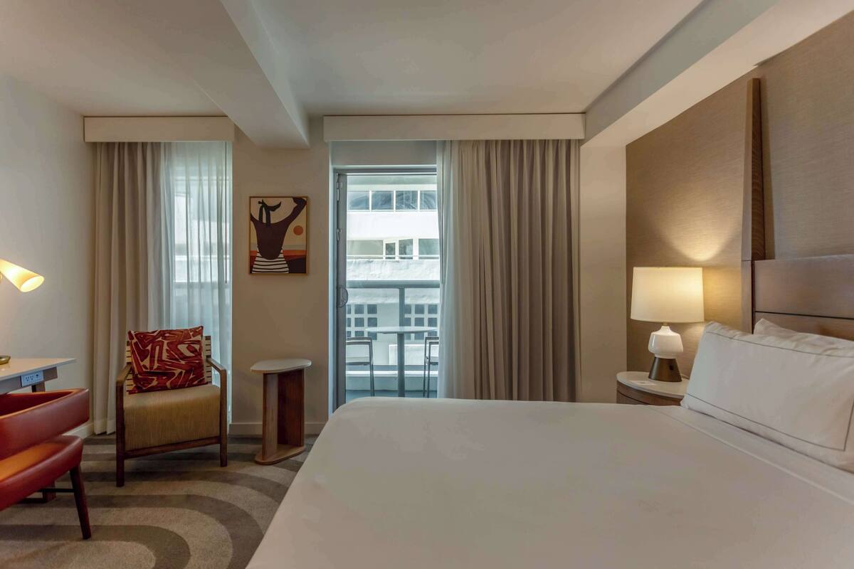 A hotel room with a large bed, a chair, a small table, and a nightstand with a lamp. There is a window with curtains and a view of a building outside.