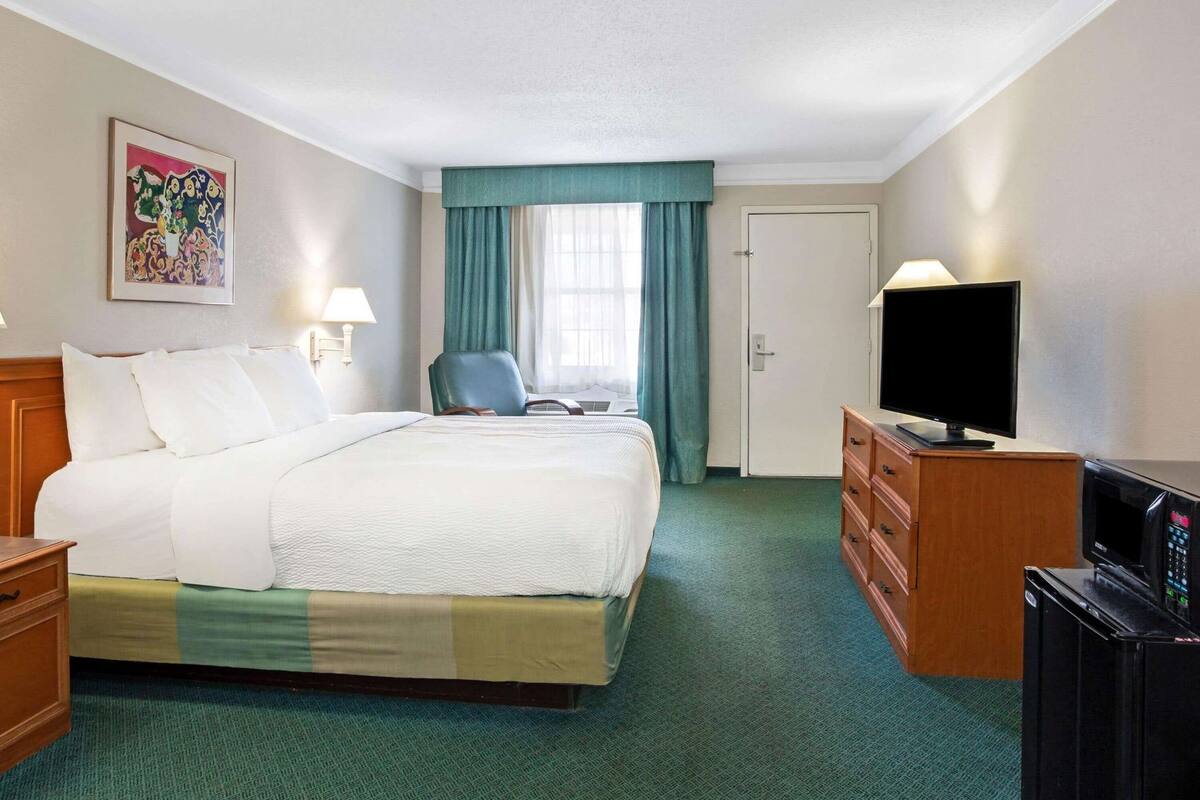 Habitación de hotel con cama, escritorio con televisor, silla, puerta y ventana con cortinas.