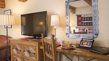 Una habitación con escritorio de madera, televisor, computadora portátil y espejo con marco decorativo.