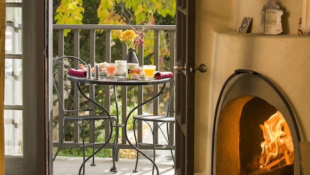 An open door leading to a balcony with a table set for two, a fireplace, and a view of trees.