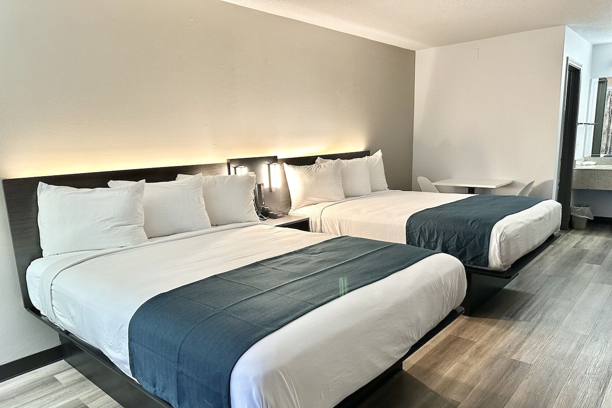A hotel room with two beds, each with white linens and dark blue bedspreads. There are bedside tables with lamps, a small desk, and a chair. The room has a wooden floor and a view of the outside.