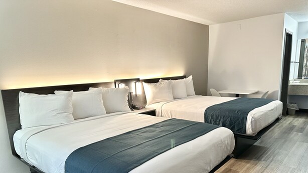 A hotel room with two beds, each with white linens and dark blue bedspreads. There is a small table and a chair in the room. - Studio 6 Suites Louisville, KY – Airport/Expo Center (Louisville)