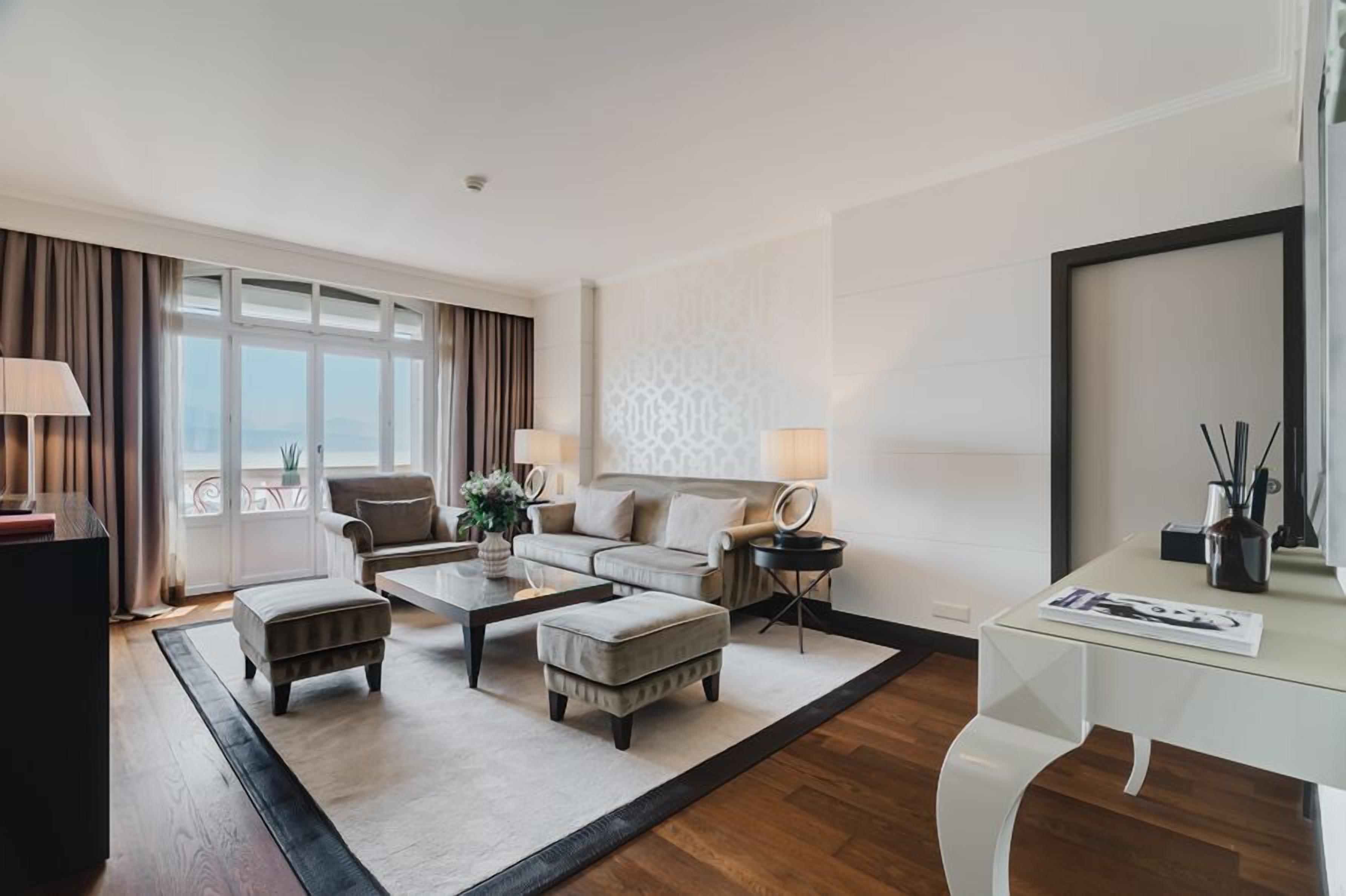Una habitación de hotel moderna con un conjunto de sofás, una mesa de centro, un escritorio y un ventanal con cortinas.