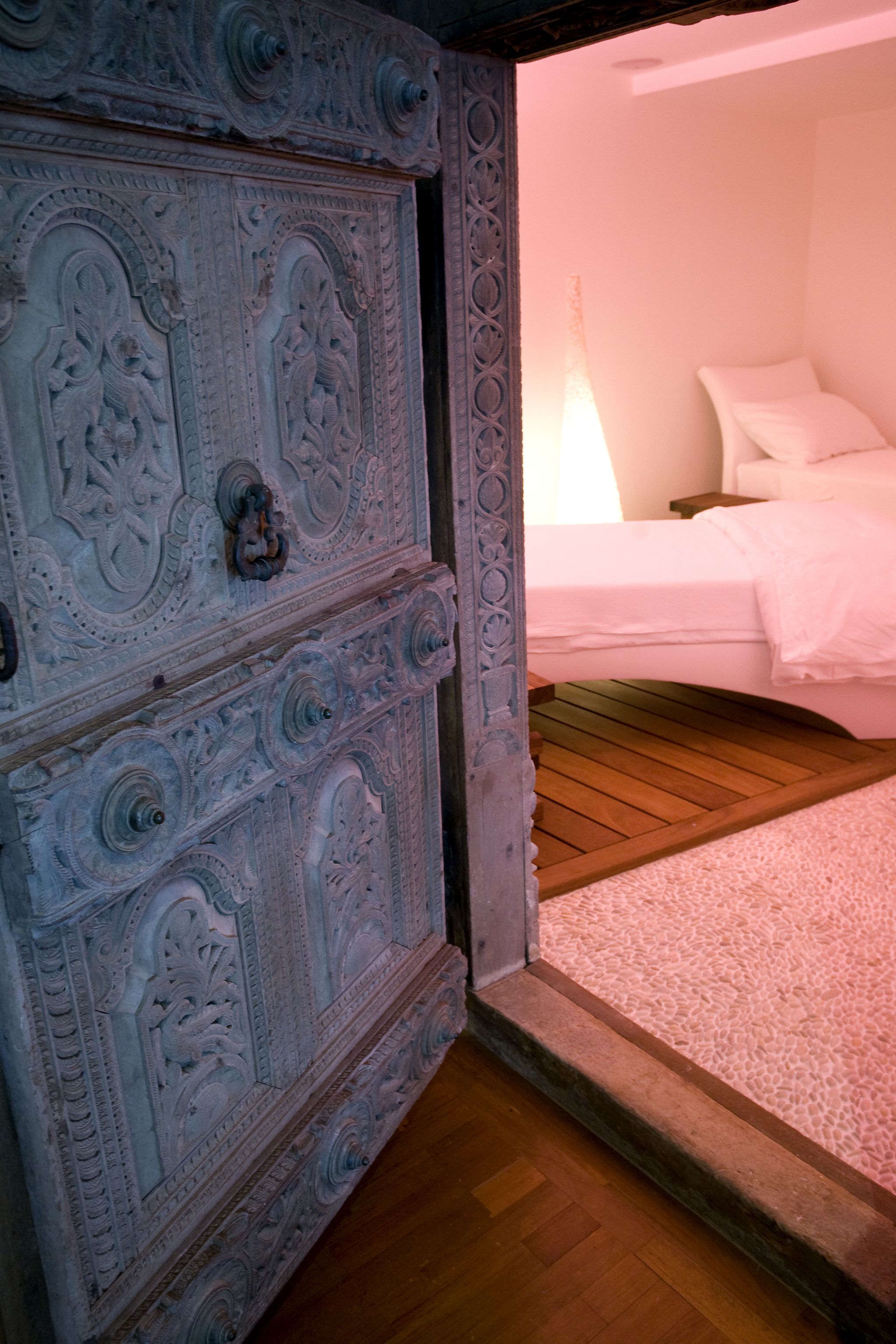 A bedroom with a wooden floor, a bed with white bedding, and a decorative door with intricate carvings.