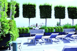 A rooftop terrace with lounge chairs, a wooden bench, and neatly trimmed hedges.