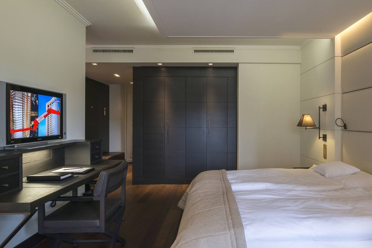 A modern hotel room with a bed, a desk with a computer, a television, and a wardrobe.
