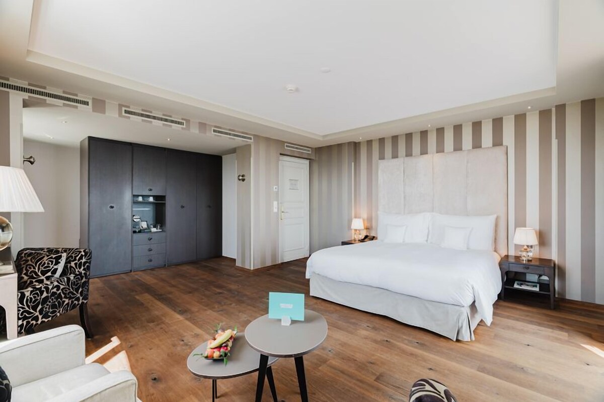A hotel room with a large bed, a desk, a chair, a small table, and a wardrobe.