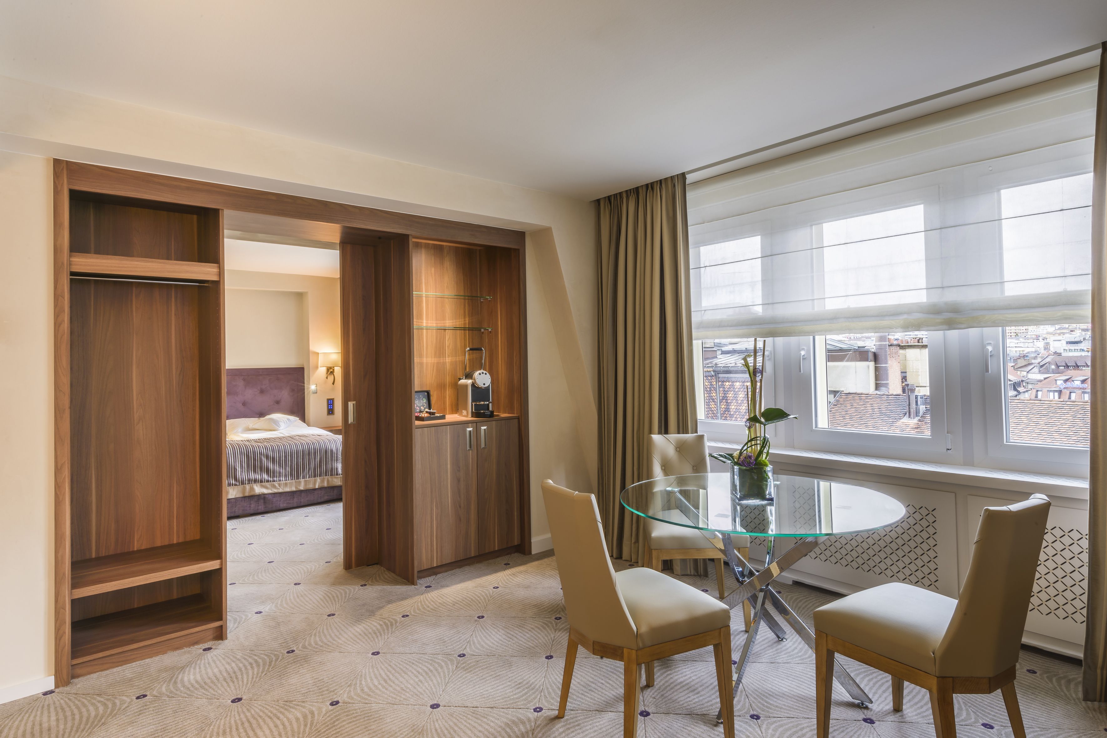 Habitación de hotel con cama, mesa de comedor de vidrio con sillas, armario de madera y vista de la ciudad a través de amplias ventanas.