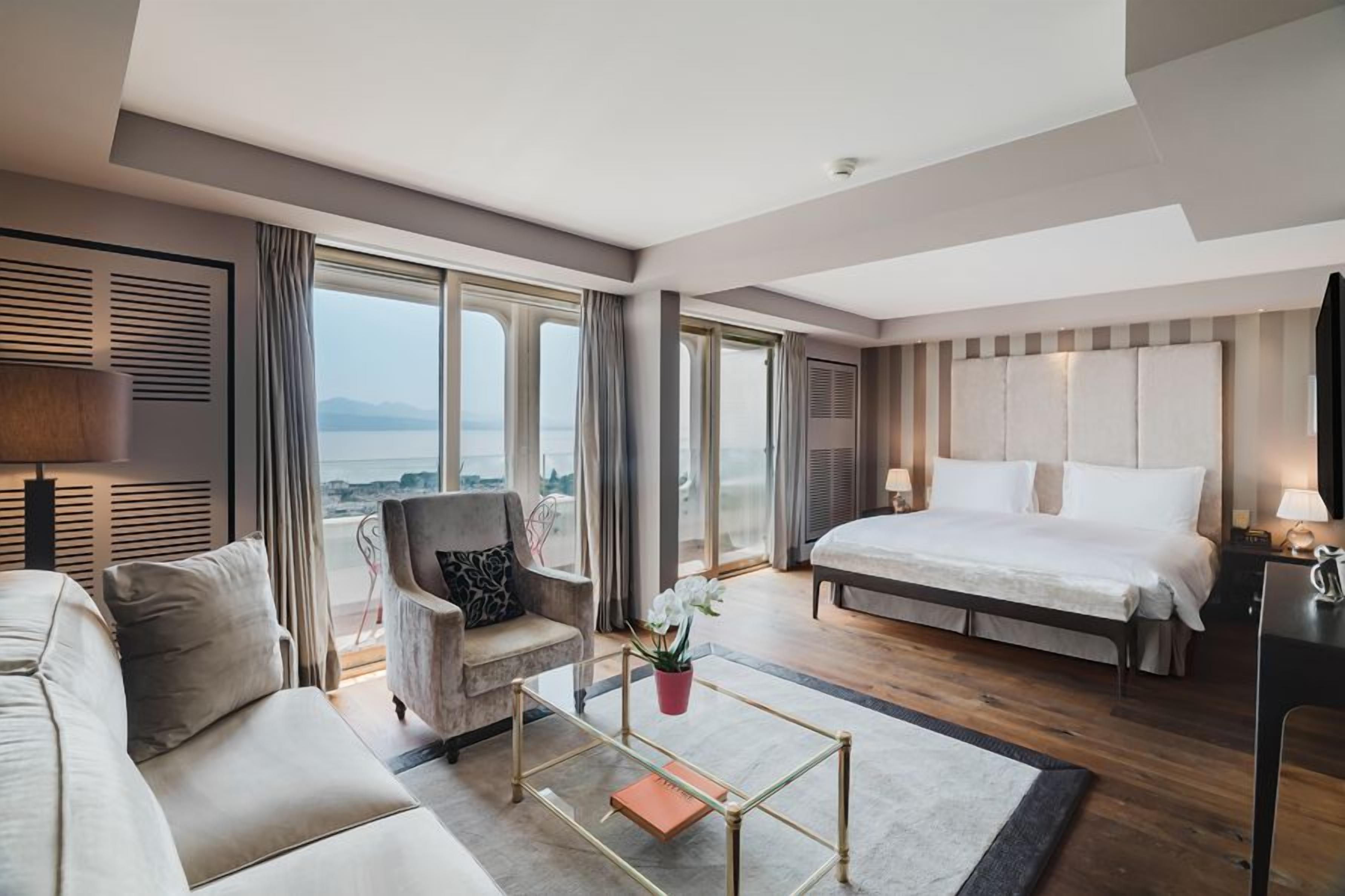 A modern hotel room with a large bed, a sofa, a chair, and a view of the sea.